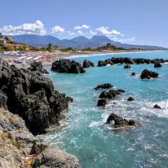 Vue d'une plage animée avec des parasols colorés, des rochers sombres et une mer turquoise. La **scalea** de la côte italienne offre un paysage pittoresque.