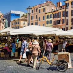 Animation d'un marché animé à Rome, avec des étals de fruits et légumes sous des parasols blancs. Une bicyclette jaune est garée près d'un chariot.