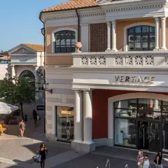Boutiques de luxe, dont Versace, dans un centre commercial de style italien près de Rome. Des clients flânent sous le soleil.