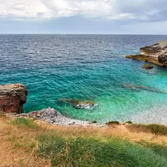 Vue d'une crique aux eaux turquoise cristallines, typique d'une plage de Pula en Sardaigne. Rochers et végétation méditerranéenne bordent la petite plage de galets.