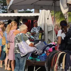 Jour de marché à Albenga : des gens flânent devant des étals de vêtements et d'accessoires, sous les palmiers.