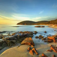 Coucher de soleil sur une plage rocheuse en Maremma, Italie. L'eau calme caresse le sable doré et les rochers ocre sous un ciel aux teintes chaudes.