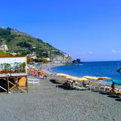 Plage de sable noir à Maiori, Italie, avec parasols, chaises longues, un catamaran blanc et des collines verdoyantes.