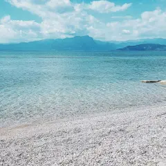 Plage de galets blancs au bord du lac de Garde, eaux bleues transparentes, montagnes bleues à l'horizon sous un ciel nuageux.