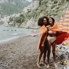 Deux amies rient sur une plage italienne, partageant une serviette orange. Leurs vacances en Italie en famille au bord de la mer sont idylliques.