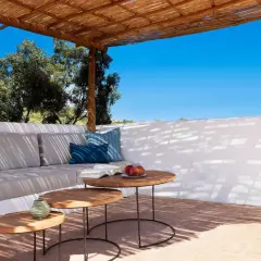 Terrasse ombragée d'un agriturismo en Sardaigne, avec canapé confortable, tables basses en bois et vue sur la végétation luxuriante sous un ciel bleu éclatant.