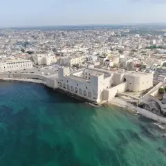 Vue aérienne du château de Trani, une forteresse médiévale sur la côte, avec la ville en arrière-plan et la mer d'un bleu-vert éclatant.