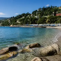 Plage de galets à Rapallo, avec des cabines bleues et blanches alignées, des maisons colorées sur la colline verdoyante et une mer bleue étincelante.