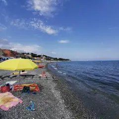 Plage de galets animée, avec des parasols, des serviettes et des baigneurs profitant du soleil. La mer bleue s'étend à l'horizon, avec des bateaux au loin. Un aperçu de la vie sur la côte ligure.