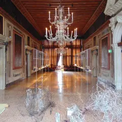 Intérieur du Palazzo Mocenigo, un long couloir orné de tableaux, avec un lustre imposant et des sculptures modernes au sol.