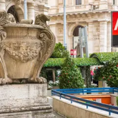 Un signe du **metro vatican** indique l'entrée d'une station, à côté d'une urne antique et de verdure luxuriante.