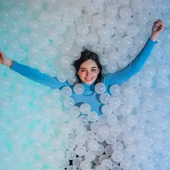 Une jeune femme souriante se détend dans une piscine de balles blanches, un clin d'œil ludique à l'ikono Rome.