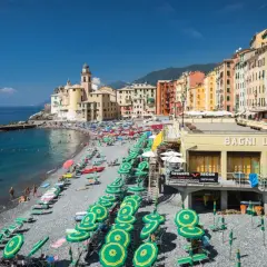 Plage animée à Camogli, Ligurie, avec des parasols verts et jaunes, des bâtiments colorés et la mer bleue.