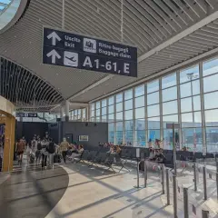 Intérieur lumineux d'un aéroport, avec des panneaux indiquant "Uscita" et "Ritiro bagagli". Des voyageurs attendent près des grandes baies vitrées, prêts pour leur vol au départ de l'aéroport Rome centre ville.
