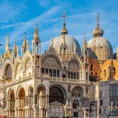 La basilique Saint-Marc à Venise, avec ses dômes majestueux et sa façade ornée, se dresse sous un ciel bleu éclatant. Un oiseau survole ce chef-d'œuvre architectural.