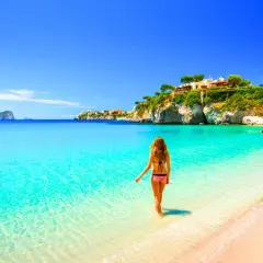Femme en bikini entrant dans l'eau turquoise d'une plage idyllique, avec des maisons perchées sur une falaise, typique de la côte méditerranée en Italie.