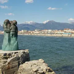 Sculpture de famille sur un rocher, face à la mer et à la ville de via Reggio, avec des montagnes enneigées en arrière-plan.