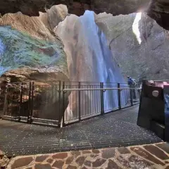 Vue depuis une grotte sur la cascade du Varone, avec une passerelle et un visiteur admirant la chute d'eau.