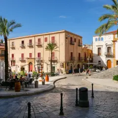 Place animée de Cefalù avec des terrasses de café, des palmiers et des bâtiments historiques sous un ciel bleu.