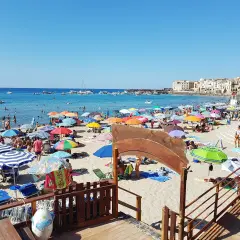 Plage animée à Cefalù en Sicile, avec des parasols colorés, des baigneurs dans la mer bleue et des bâtiments historiques en arrière-plan.