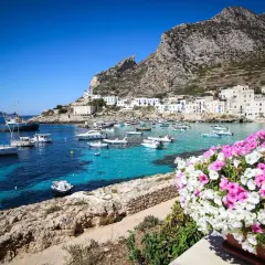 Vue d'un port pittoresque en Italie, avec des bateaux dans des eaux turquoise et des maisons blanches accrochées à flanc de colline. Un bouquet de fleurs roses et blanches encadre la scène, évoquant la beauté de Marsala, Italie.