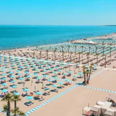 Plage de Chioggia, Italie, avec des rangées infinies de parasols bleus et blancs, des chaises longues et des palmiers sous un ciel bleu éclatant.