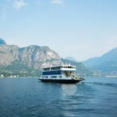 Un ferry transporte des voitures sur le lac de Côme, naviguant paisiblement entre des montagnes majestueuses sous un ciel bleu.