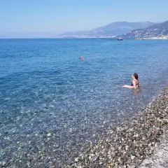 Une femme se baigne dans les eaux claires de la plage de galets de Ventimiglia, avec des montagnes en arrière-plan.