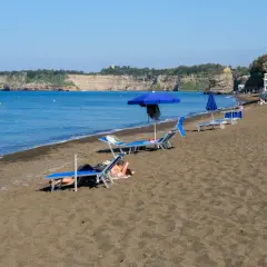 Plage de sable noir avec des chaises longues et des parasols bleus, face à une falaise rocheuse. Un doux air de Procida ou Capri.