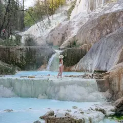 Une femme en maillot de bain blanc et chapeau de paille admire les cascades et les bassins turquoise des Bagni San Filippo, un paysage naturel spectaculaire.