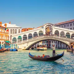 Gondole naviguant sur le Grand Canal, sous le célèbre pont du Rialto à Venise. L'eau turquoise reflète les façades colorées des bâtiments.