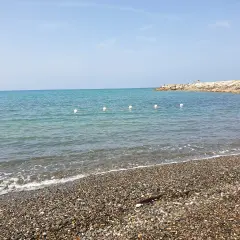 Plage de galets à Cecina Mare, avec des bouées blanches marquant la zone de baignade et une jetée rocheuse au loin.