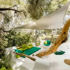 Terrasse du Hotel Su Gologone avec mobilier blanc et coussins verts, sous une voile d'ombrage et entourée de verdure.