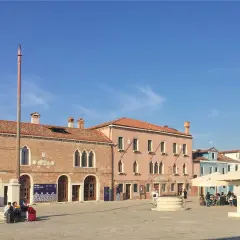 Place du **musée de la dentelle Burano**. Bâtiments colorés, mât, puits et statue sous un ciel bleu.