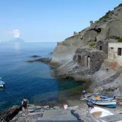 Sur l'île Salina, un homme amarre un bateau près de vieilles maisons de pêcheurs en pierre, avec une île volcanique au loin.