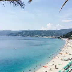 Plage animée de la riviera italienne, avec des parasols verts, des baigneurs dans la mer bleue et des collines verdoyantes en arrière-plan.