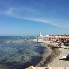 Plage animée à San Cataldo, Pouilles, avec des parasols orange, des baigneurs sur un ponton et un phare blanc en arrière-plan.