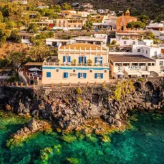 Vue d'un gîte en Sicile, perché sur une falaise rocheuse surplombant une mer turquoise. Bâtiments blancs et ocres, végétation luxuriante.