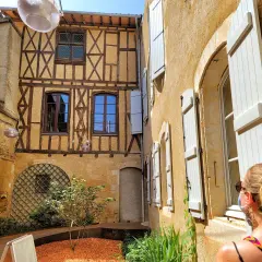 Cour intérieure avec colombages et volets blancs. Une femme regarde la scène, comme si elle découvrait un coin charmant de Fleurance, Italie.