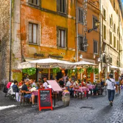 Une trattoria animée à Rome, avec des tables dressées sur la rue pavée. Des gens savourent un repas sous les parasols, profitant de l'ambiance romaine.