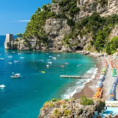 Vue panoramique d'une plage de galets animée à Almafi, Italie, avec des parasols colorés, des chaises longues et des eaux bleues claires parsemées de bateaux.