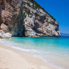 Plage de sable fin bordée par une eau turquoise cristalline, avec une falaise imposante en arrière-plan. Un coin de paradis sur la côte smeralda sardaigne.