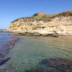 Une femme en bikini profite de la mer limpide près d'une falaise rocheuse à Anzio, avec une tour ancienne en arrière-plan.