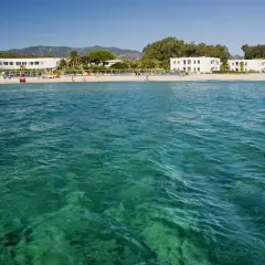 Eaux turquoise cristallines devant une plage de sable fin à Pula, Sardaigne, avec des hôtels blancs et des palmiers.