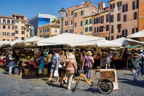 Animation d'un marché animé à Rome, avec des étals de fruits et légumes sous des parasols blancs. Une bicyclette jaune est garée près d'un chariot.