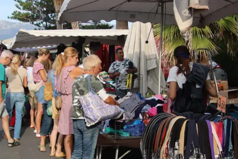 Jour de marché à Albenga : des gens flânent devant des étals de vêtements et d'accessoires, sous les palmiers.