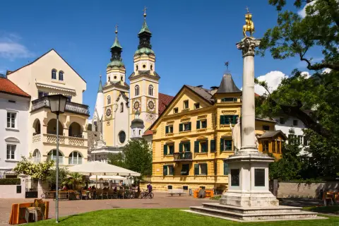 Place ensoleillée à Bressanone avec des bâtiments colorés, une colonne monumentale et des terrasses de café animées.