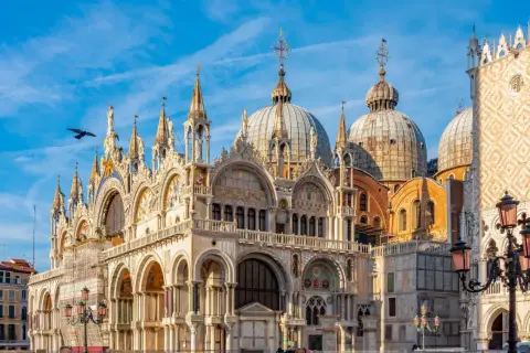 La basilique Saint-Marc à Venise, avec ses dômes majestueux et sa façade ornée, se dresse sous un ciel bleu éclatant. Un oiseau survole ce chef-d'œuvre architectural.