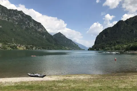 Un kayak attend sur la rive du lac d'Idro, entouré de montagnes verdoyantes sous un ciel bleu parsemé de nuages.