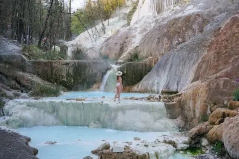 Une femme en maillot de bain blanc et chapeau de paille admire les cascades et les bassins turquoise des Bagni San Filippo, un paysage naturel spectaculaire.